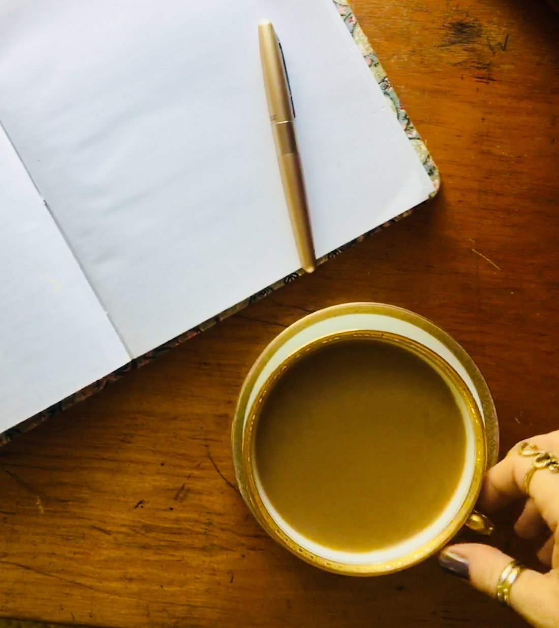 An open planning journal with a gold pen, a gilt-rimmed teacup and a hand wearing gold rings on a dark wood table